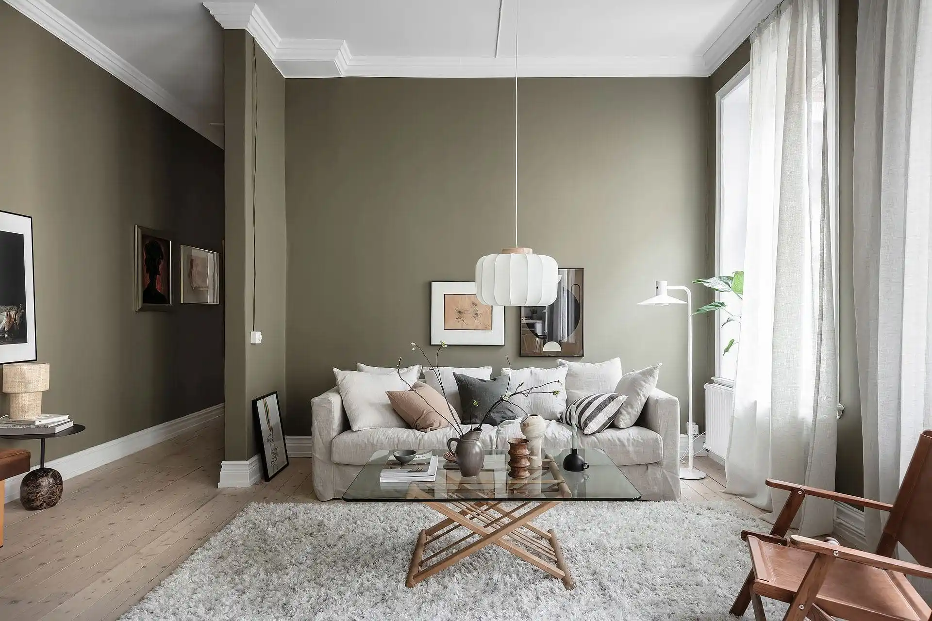 Naast kussens in hetzelfde materiaal en kleur als de bank, zijn er ook kussens gebruikt in andere kleurtinten, die complementair zijn aan de beige bank en olijfgroene muur.